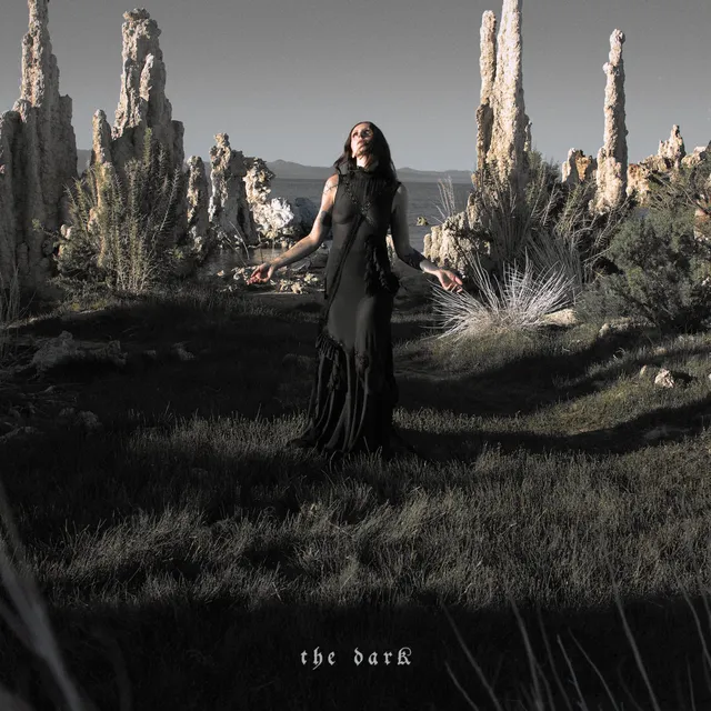 Pochette de l'album The Dark de Chelsea Wolfe : la musicienne en robe noire, bras ouverts, au milieu de formations rocheuses désertiques au bord d'un lac