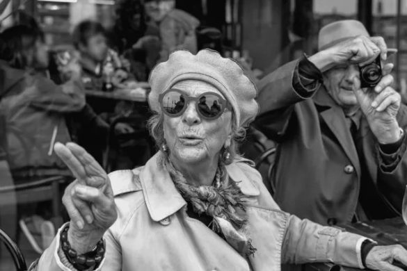 Photographie noir et blanc de Nikos Aliagas : une femme âgée au béret et lunettes rondes attablée en terrasse, un homme âgé la prend en photo derrière elle