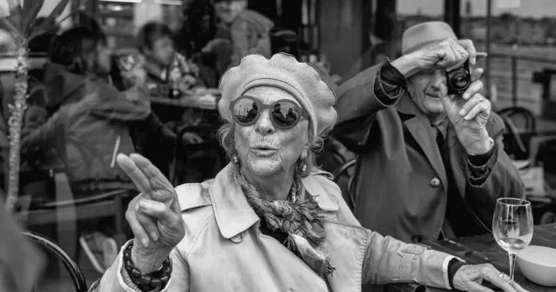 Photographie noir et blanc de Nikos Aliagas : une femme âgée au béret et lunettes rondes attablée en terrasse, un homme âgé la prend en photo derrière elle